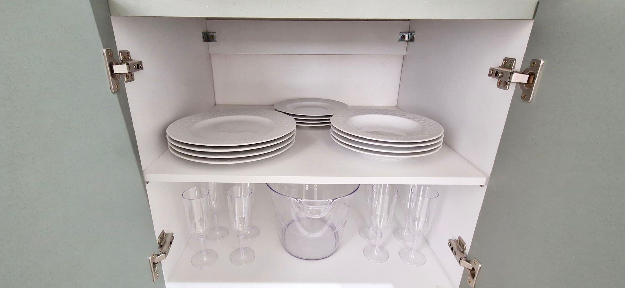 A white cabinet with two shelves; the top shelf holds stacked plates, and the bottom shelf contains wine glasses and a pitcher.
