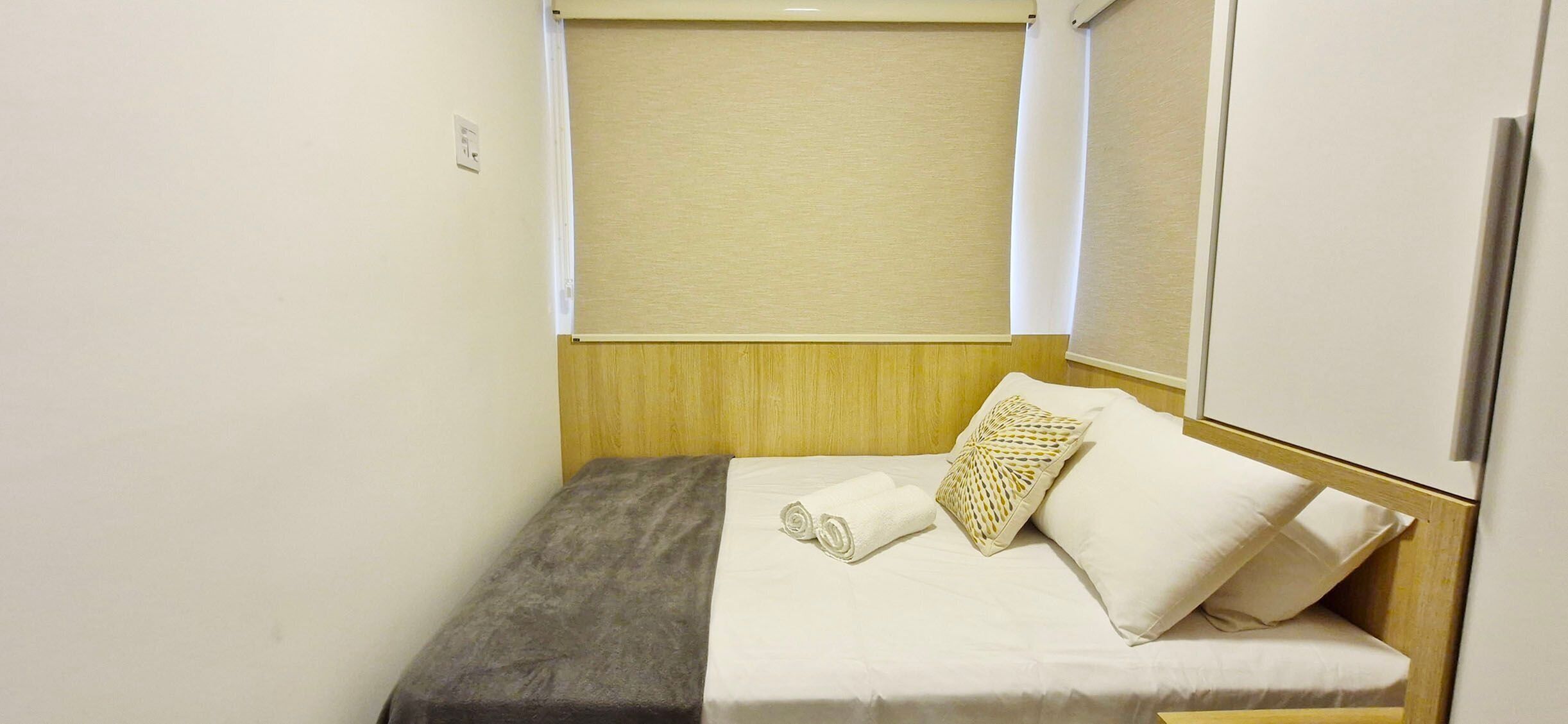 A small, single-bed room with a wooden headboard, a window with a beige blind, and a white wardrobe.