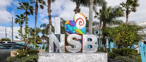 Letrero con las letras 'NSB' y un diseño de corazón colorido, rodeado de palmeras y plantas en macetas.