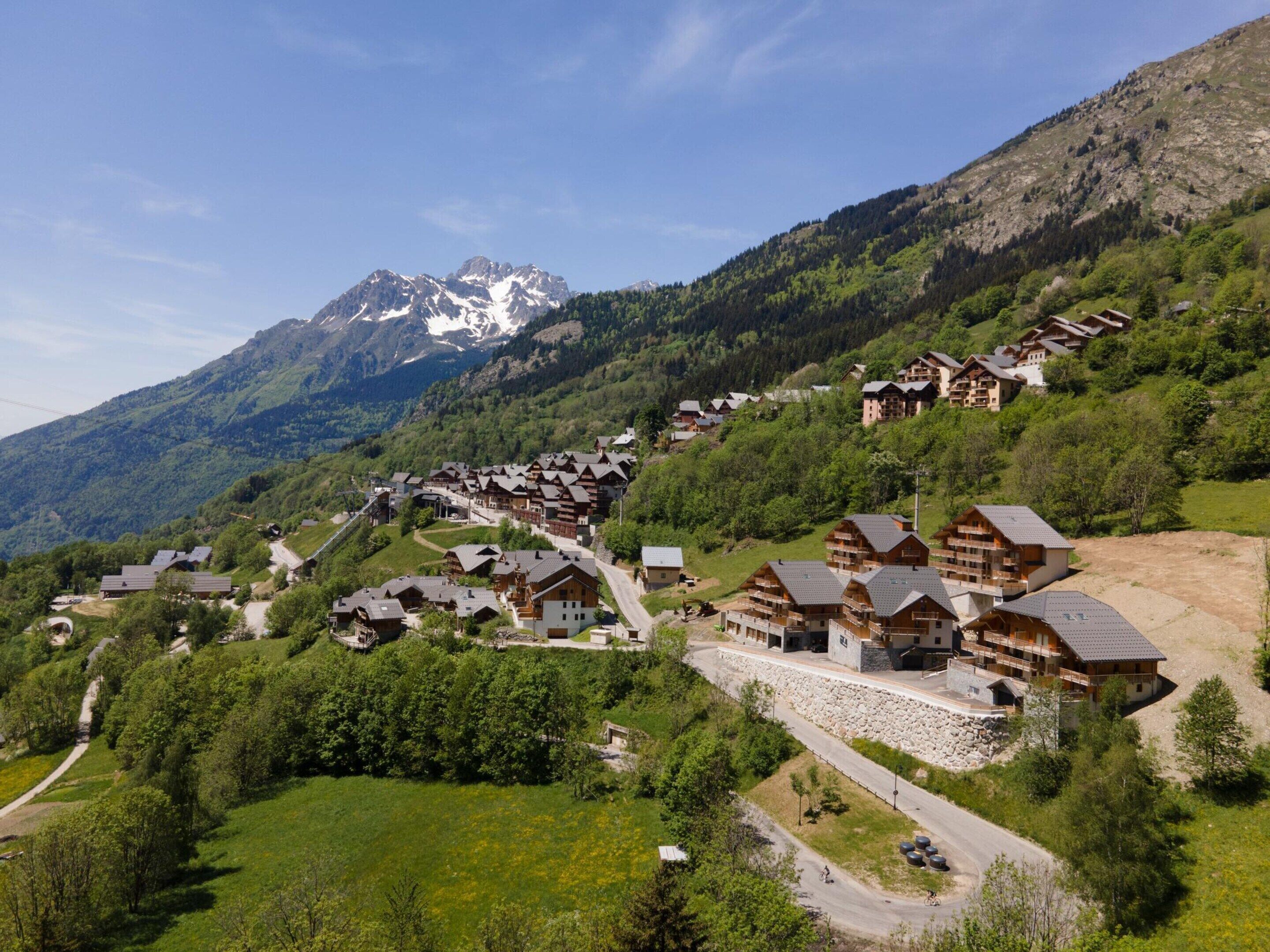 A mountain village with chalet-style buildings, a winding road, and a prominent mountain peak in the background.