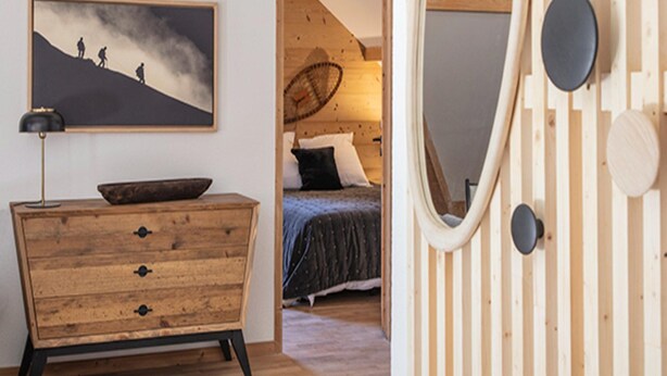 Een slaapkamer met een houten dressoir, een bed met een donkerblauwe spreideken, een grote ovale spiegel en een schilderij van een berglandschap.