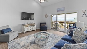 A living room with a glass coffee table, a sofa, a TV, and a view of the ocean through large sliding doors. - Charming 2-bedroom condo with fitness room, WiFi in awesome Surf City (Surf City)