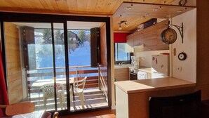 Un accogliente interno di una baita con soffitto in legno, una porta scorrevole in vetro che dà su un balcone e un angolo cottura con elettrodomestici essenziali.