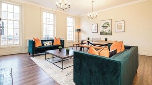 Un salon spacieux avec un canapé vert, une table basse et un coin repas avec des bancs. De grandes fenêtres et un lustre décorent la pièce.