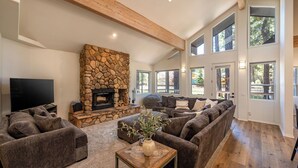 A spacious living room with a stone fireplace, large windows, and a high ceiling with exposed beams.