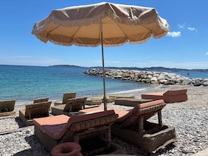 A beachside lounge with wicker furniture and a beige umbrella, overlooking a calm sea and rocky shoreline.