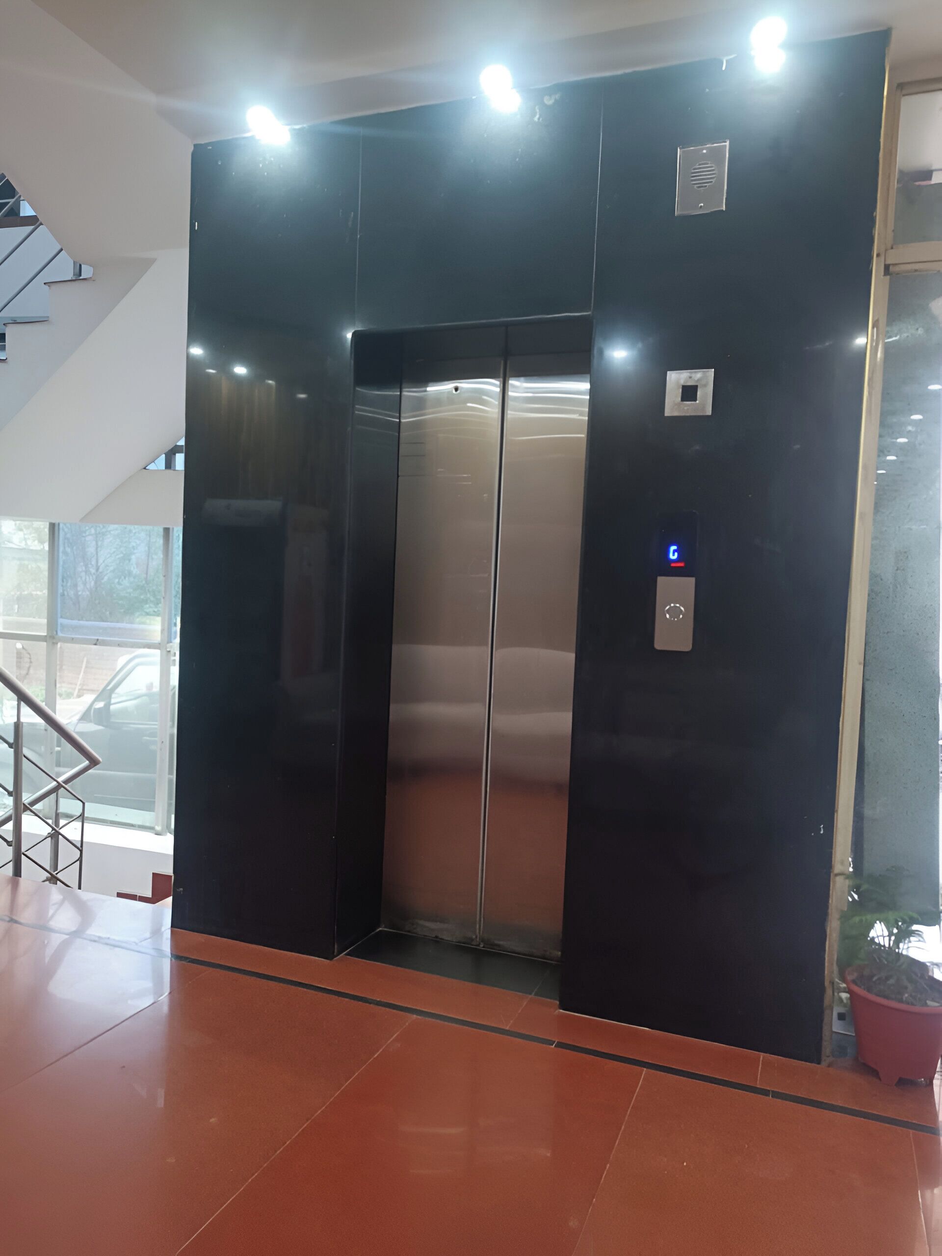 A modern elevator with a digital display, located in a lobby with a tiled floor and a staircase.