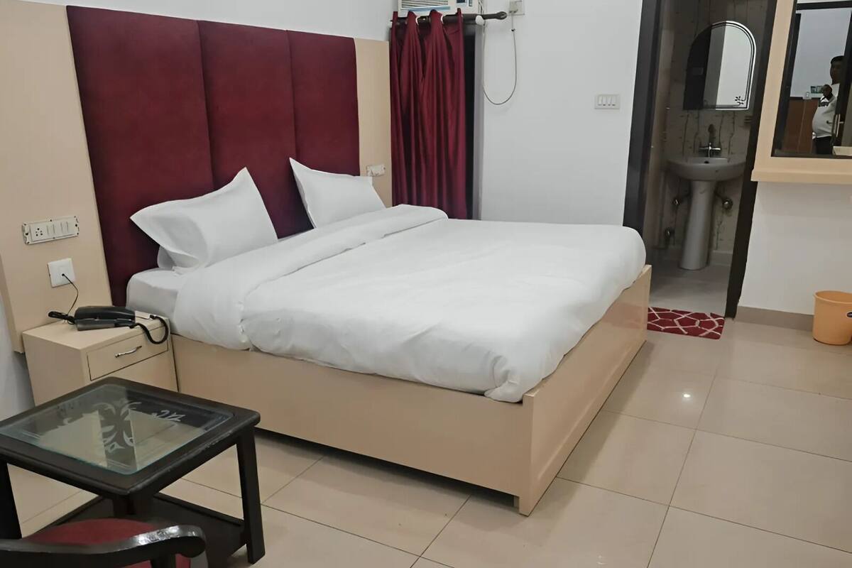 A hotel room with a bed, a bedside table with a phone, a glass table with a chair, a bathroom with a sink and mirror, and a red curtain.