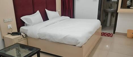 Habitación de hotel con cama, mesita de noche con teléfono, una mesa de vidrio con una silla, baño con lavamanos y espejo, y una cortina roja.