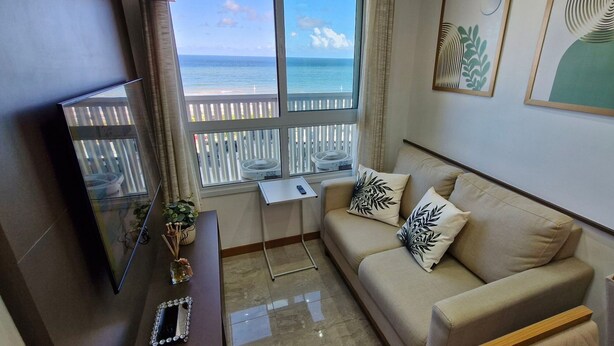 A living room with a beige sofa, two decorative pillows, a small table, a mirror, and a window with a view of the sea. - COCAZ0106 - Smart Sol do Costa Azul (Salvador)