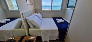 A room with two beds, each with white linens and a blue bedspread. There are two bedside tables with electronic devices. The room has large windows offering a view of the sea. - COCAZ0106 - Smart Sol do Costa Azul (Salvador)