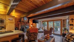 A cozy cabin interior with a wooden ceiling, a kitchen with a microwave, a bar area, a living room with a sofa and a coffee table, and a large window overlooking greenery. - Secluded Cabin w/ Spa & Games! (Big Bear)