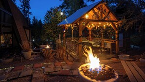 A wooden gazebo with a fire pit, surrounded by outdoor furniture and string lights. - Secluded Cabin w/ Spa & Games! (Big Bear)