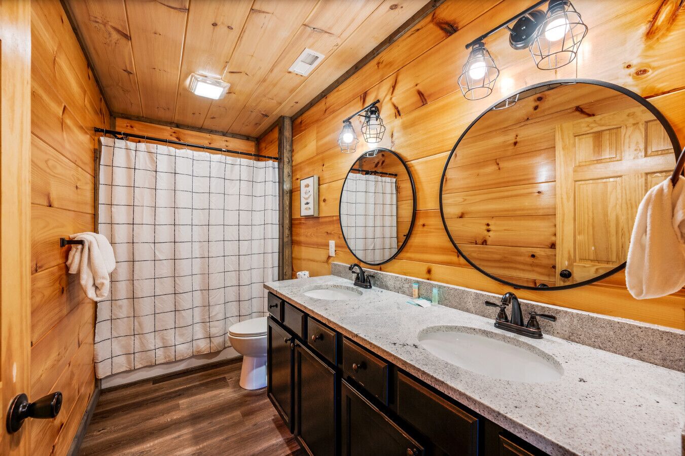 Une salle de bain rustique, avec des murs en bois, comprenant un lavabo double, deux miroirs, des toilettes et un rideau de douche.