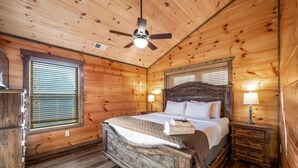 Ein Schlafzimmer einer Holzhütte mit Bett, Nachttisch, Deckenventilator und Fenster mit Jalousien.