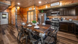 Ein gemütliches Blockhaus-Interieur mit einem Holzespeisesaal, gedeckt für sechs Personen, einer Küche mit modernen Geräten und einer Treppe, die zu einer weiteren Ebene führt.