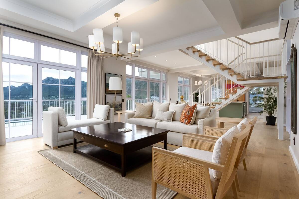 A spacious living area with a large sofa set, a coffee table, and a rattan chair. A staircase with a railing leads to an upper level.