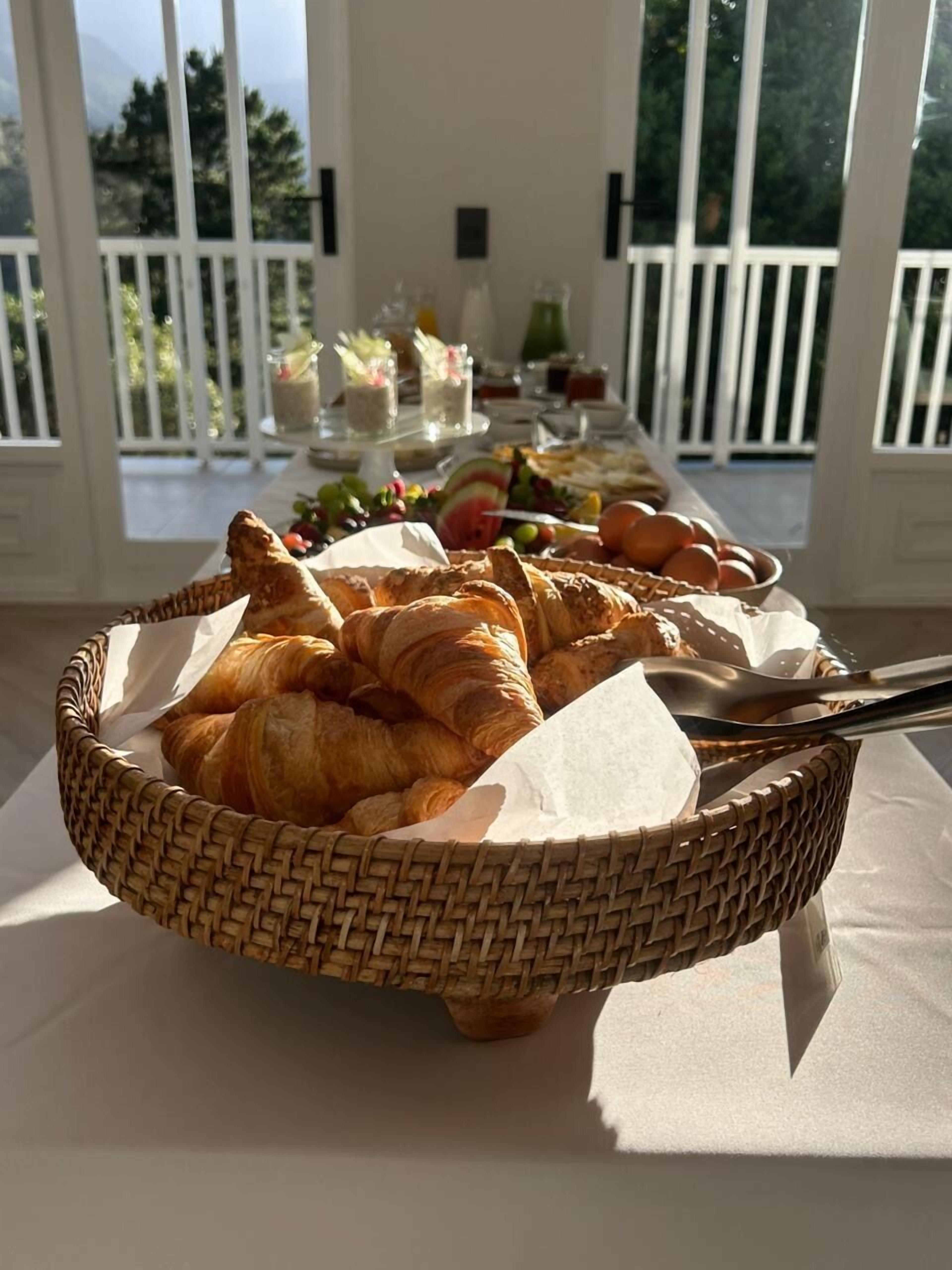 A breakfast buffet with croissants and fruits, set on a table with a view of a balcony.