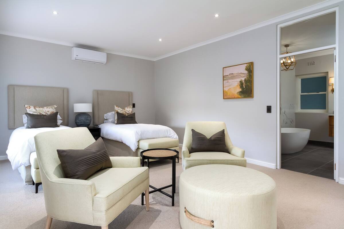 A modern hotel room with two beds, a sitting area with chairs, a round ottoman, and a view into the bathroom.