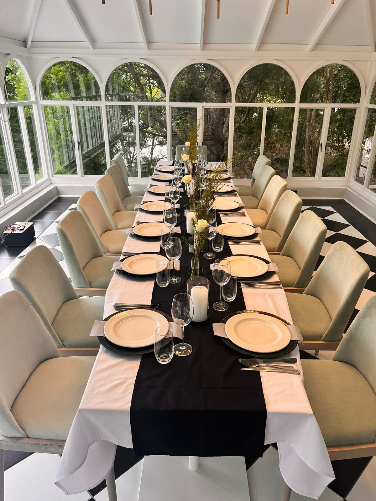 A long dining table set for a formal meal with white and black tablecloths, beige chairs, and a view of greenery through large windows.