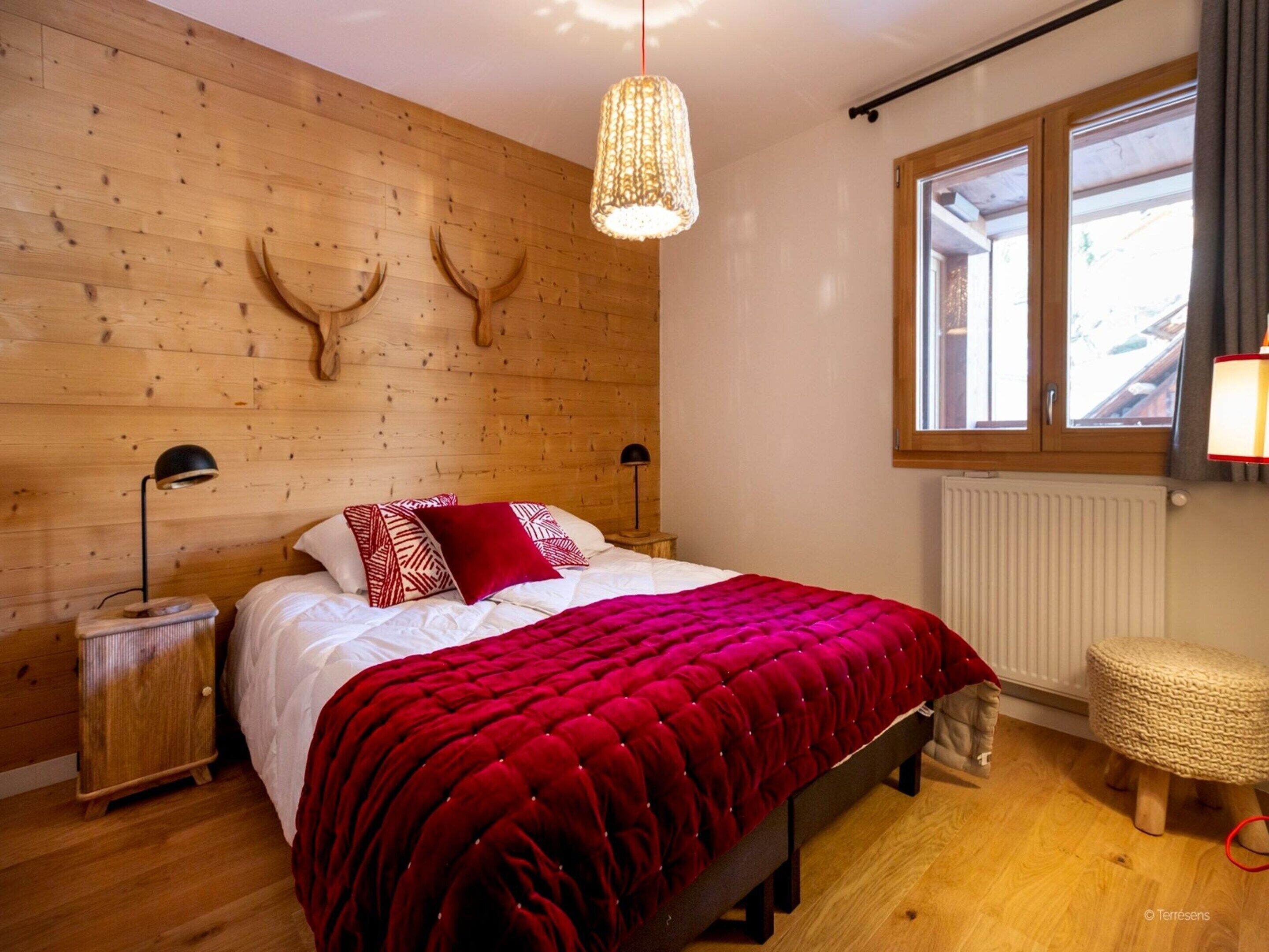 Een slaapkamer met een houten wand, een bed met een rode deken, twee bedlampjes en een raam met gordijnen.