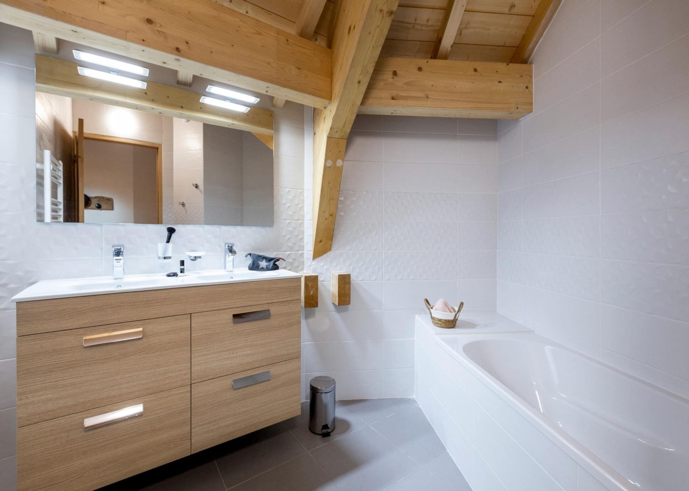 A modern bathroom with a wooden vanity, a large mirror, a bathtub, and a small wicker basket.