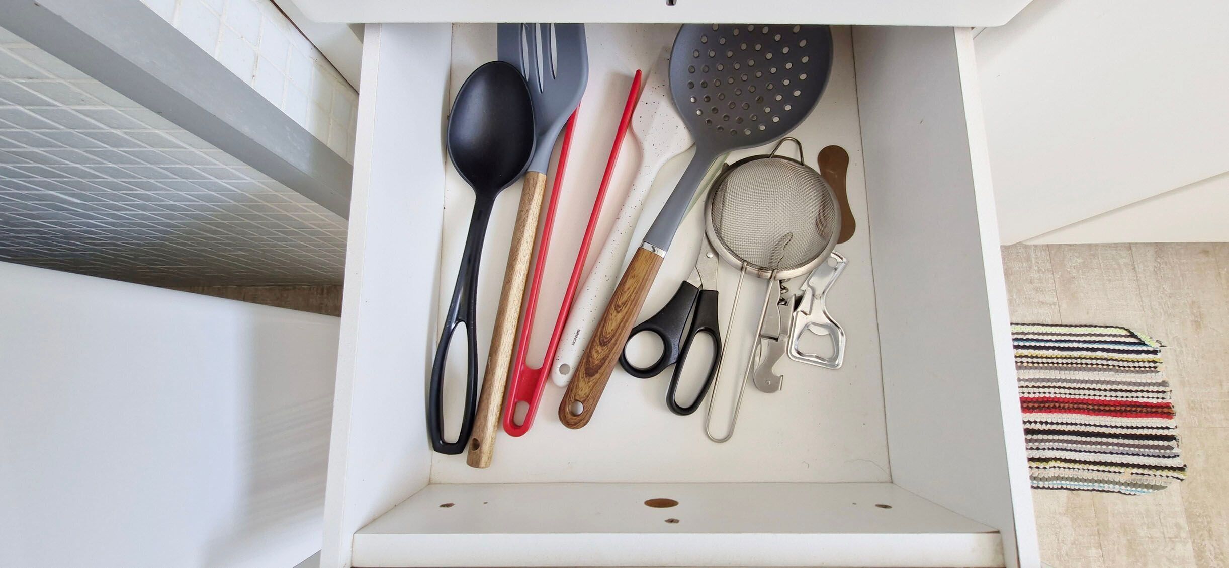 A kitchen drawer with various utensils including a black spoon, red spatula, and a metal strainer.