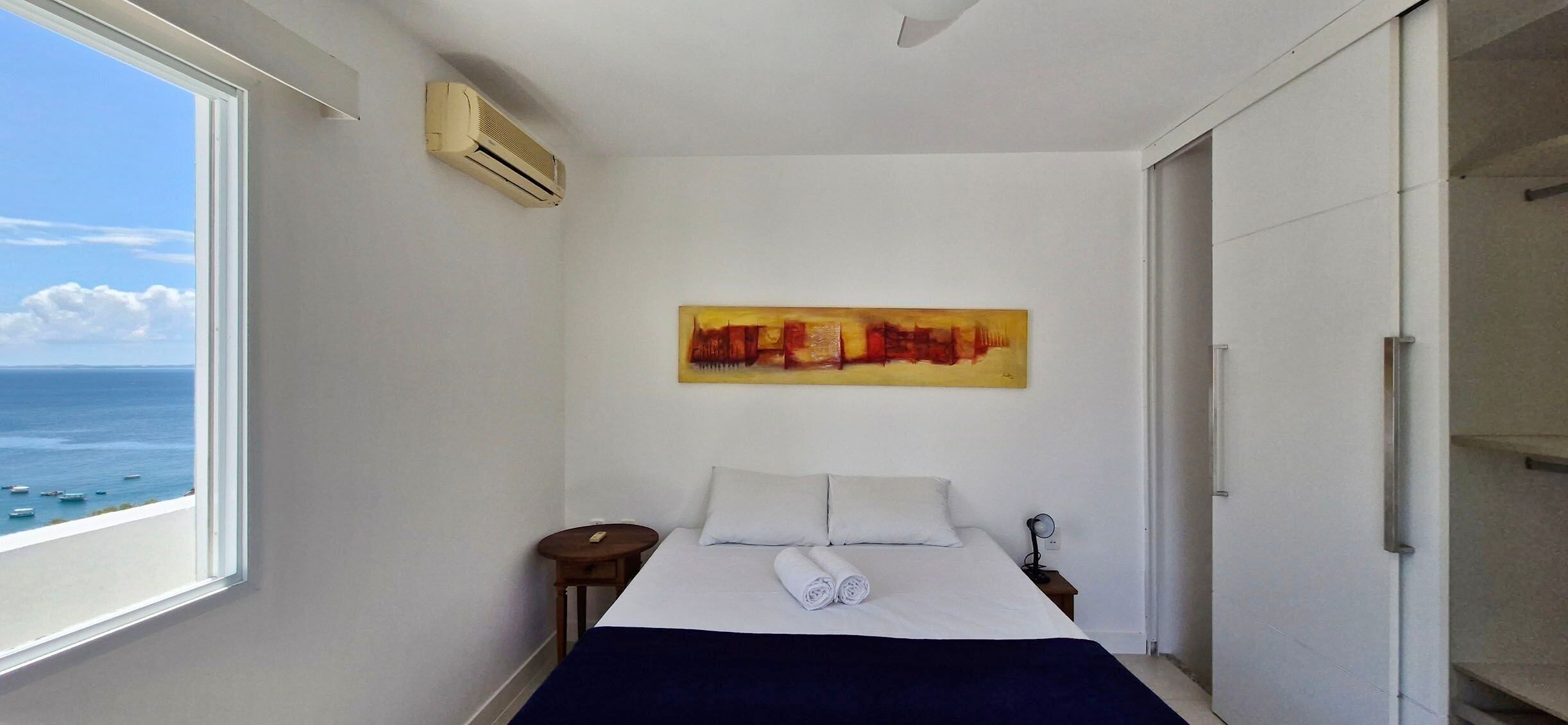 A bedroom with a bed, a painting, a bedside table, and a view of the sea.