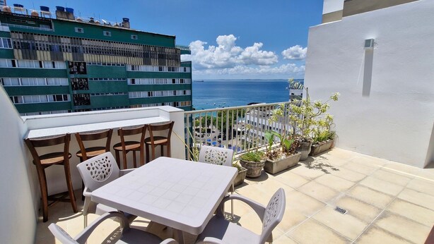 A balcony with a table and chairs, potted plants, and a view of the sea. - COBAR0130 - Edifício Porto do Sol (Salvador)