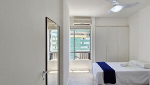 A bedroom with a bed, a wardrobe, a ceiling fan, and a window with a view of buildings. - COBAR0130 - Edifício Porto do Sol (Salvador)
