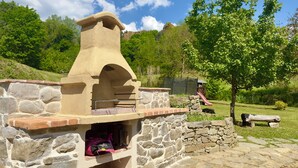 A stone barbecue grill with a chimney, set against a backdrop of trees and a playground. - Milu's Holiday Home (Toscana)