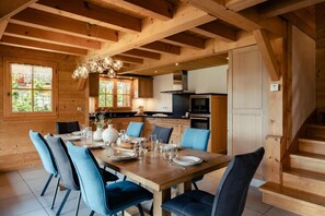 Ein Holzinterieur mit einem Esstisch, gedeckt für sechs Personen, blau gepolsterten Stühlen, einer Chandelieber und einer Küchenzeile mit modernen Geräten.