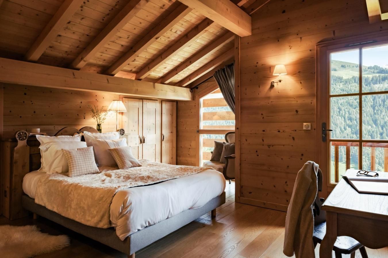 A wooden cabin bedroom with a bed, bedside lamps, a view of the forest through a window, and a table with a chair.