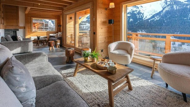 A cozy wooden interior with a sofa, armchairs, and a coffee table. Large windows offer a view of snow-capped mountains.