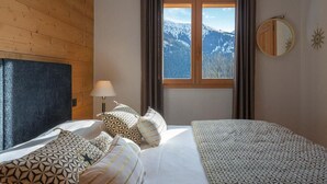 A bedroom with a bed, pillows, a nightstand, a lamp, a window with curtains, a mirror, and a view of mountains.