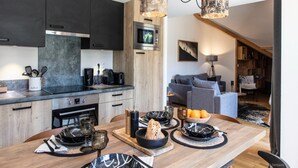 A modern kitchen with dark cabinets, a wooden dining table set with plates, glasses, and a bowl of fruit. - Premium Apartment in Vaujany (Vaujany)