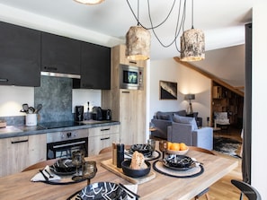 A modern kitchen with dark cabinets, a wooden dining table set with plates, glasses, and a bowl of fruit. - Premium Apartment in Vaujany (Vaujany)