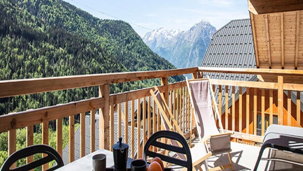 A wooden deck with a table and chairs, overlooking a forested mountain range. - Premium Apartment in Vaujany (Vaujany)