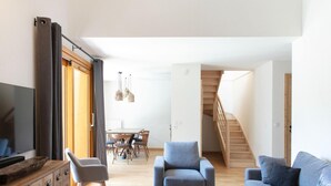 A modern living room with a sofa, armchairs, a coffee table, and a TV. There's a dining area with a table and chairs, and a staircase leading upstairs. - Premium Apartment in Vaujany (Vaujany)
