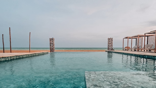 Une piscine aux eaux cristallines, entourée d’une terrasse carrelée, avec vue sur la plage, et une structure en bois agrémentée de chaises longues.