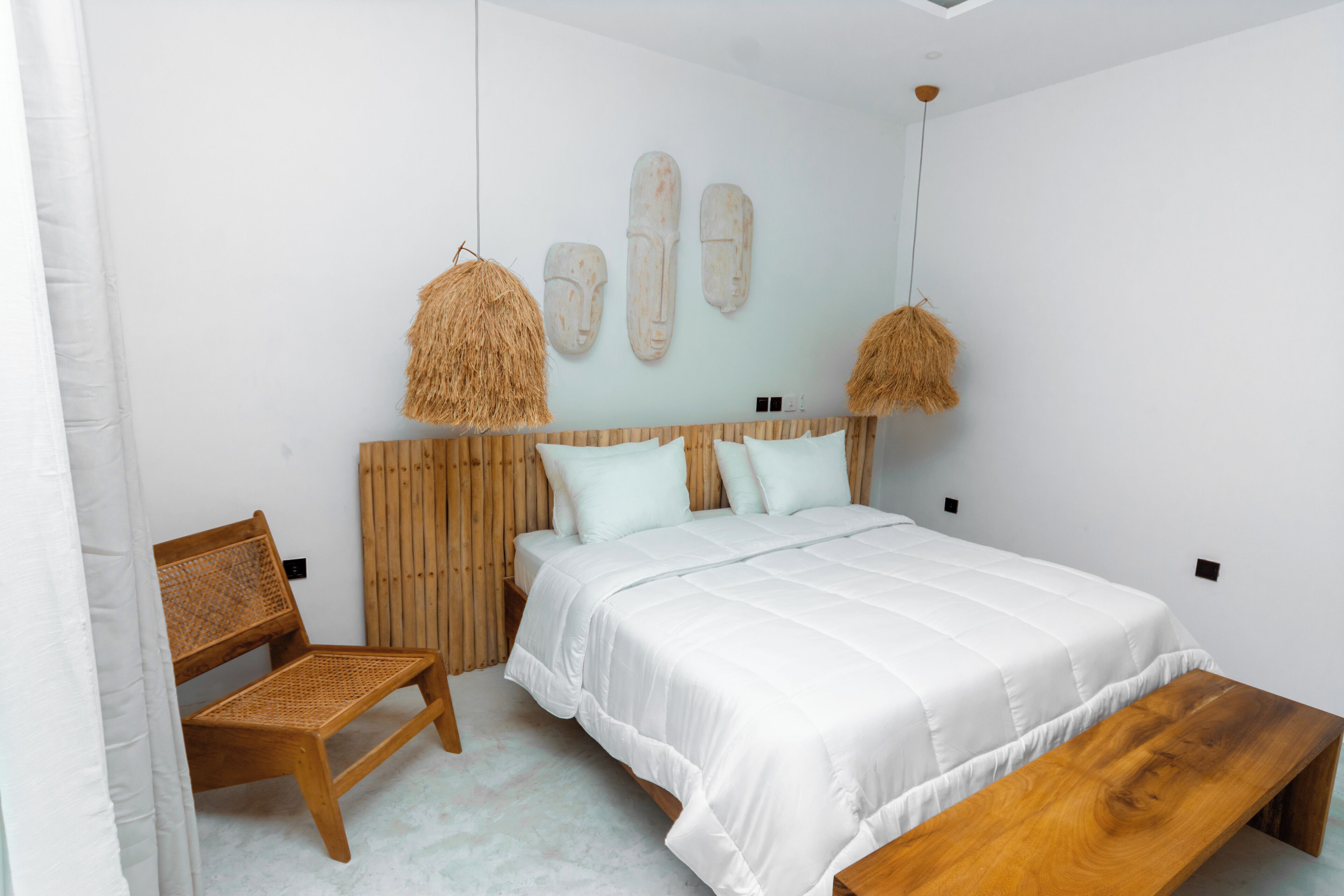 Un dormitorio con cama, un banco de madera, una silla de mimbre y un cabecero fijado a la pared.