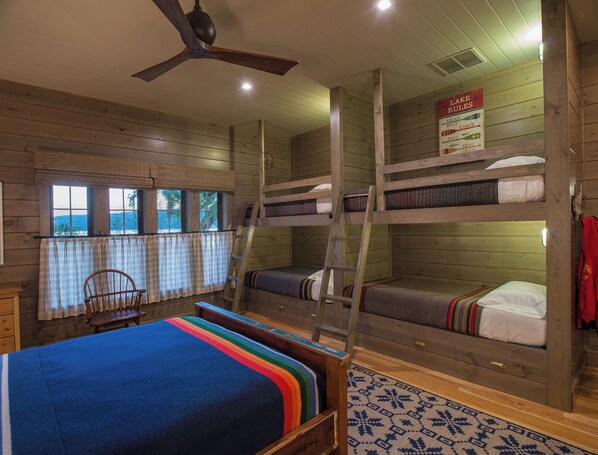 A cabin-style room with bunk beds, a ceiling fan, a window with curtains, and a sign listing room rules.