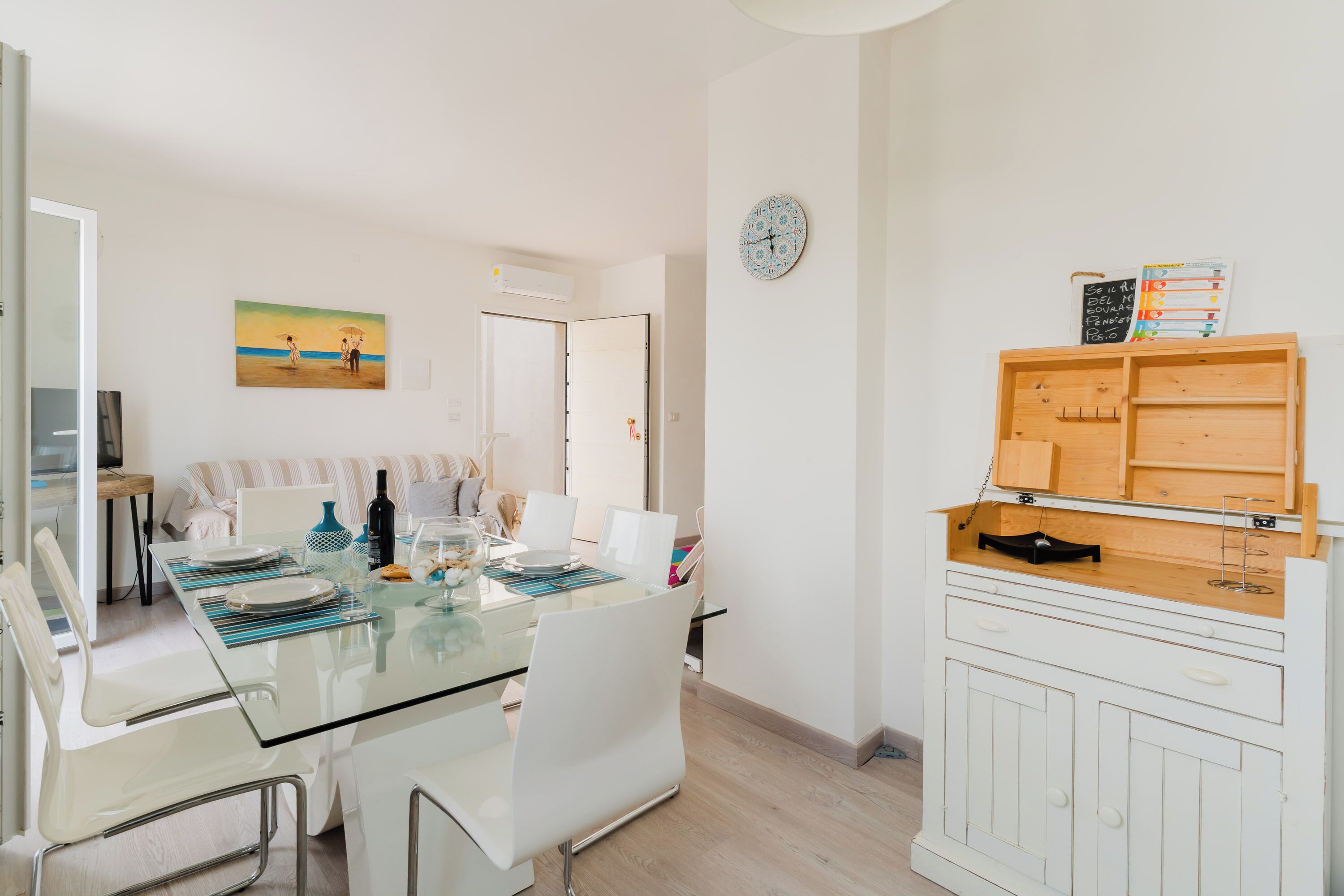 A modern dining area with a glass table, white chairs, and a wooden cabinet with a clock and educational materials.