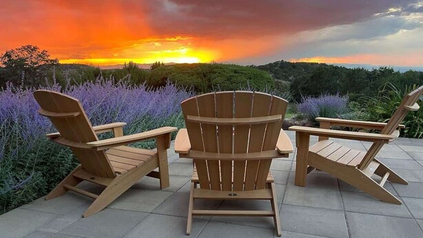 Three wooden Adirondack chairs and a table on a patio with a sunset view.