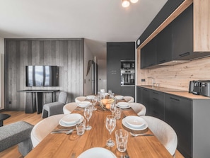 A modern dining area with a wooden table set for six, surrounded by dark cabinetry and a kitchen area with a microwave and oven. - Apartment in Plan Peisey Near Paradiski (Peisey-Nancroix)