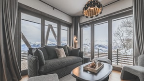 A modern living room with a sofa, ottoman, and coffee table, large windows with a mountain view, and a unique pendant light. - Apartment in Plan Peisey Near Paradiski (Peisey-Nancroix)
