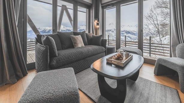 A modern living room with a sofa, ottoman, and coffee table, large windows with a mountain view, and a unique ceiling light fixture. - Apartment in Plan Peisey Near Paradiski (Peisey-Nancroix)