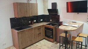 Cocina compacta con armarios de madera, encimera negra, horno empotrado, televisor de pared y un pequeño comedor con taburetes.