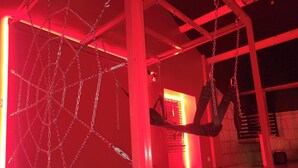 Una habitación con ambiente rojizo, que presenta una estructura metálica colgante que recuerda a una tela de araña y un arnés de cuero negro suspendido.