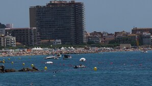 A coastal cityscape with a large hotel building, a busy beach with people and boats, and clear skies. - noo............................. (Sant Antoni de Calonge)